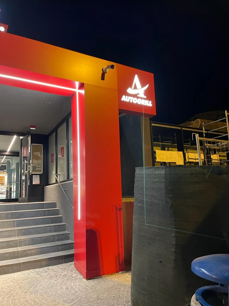 Dettaglio della spalla rossa del portale con logo Autogrill e linea luminosa verticale.