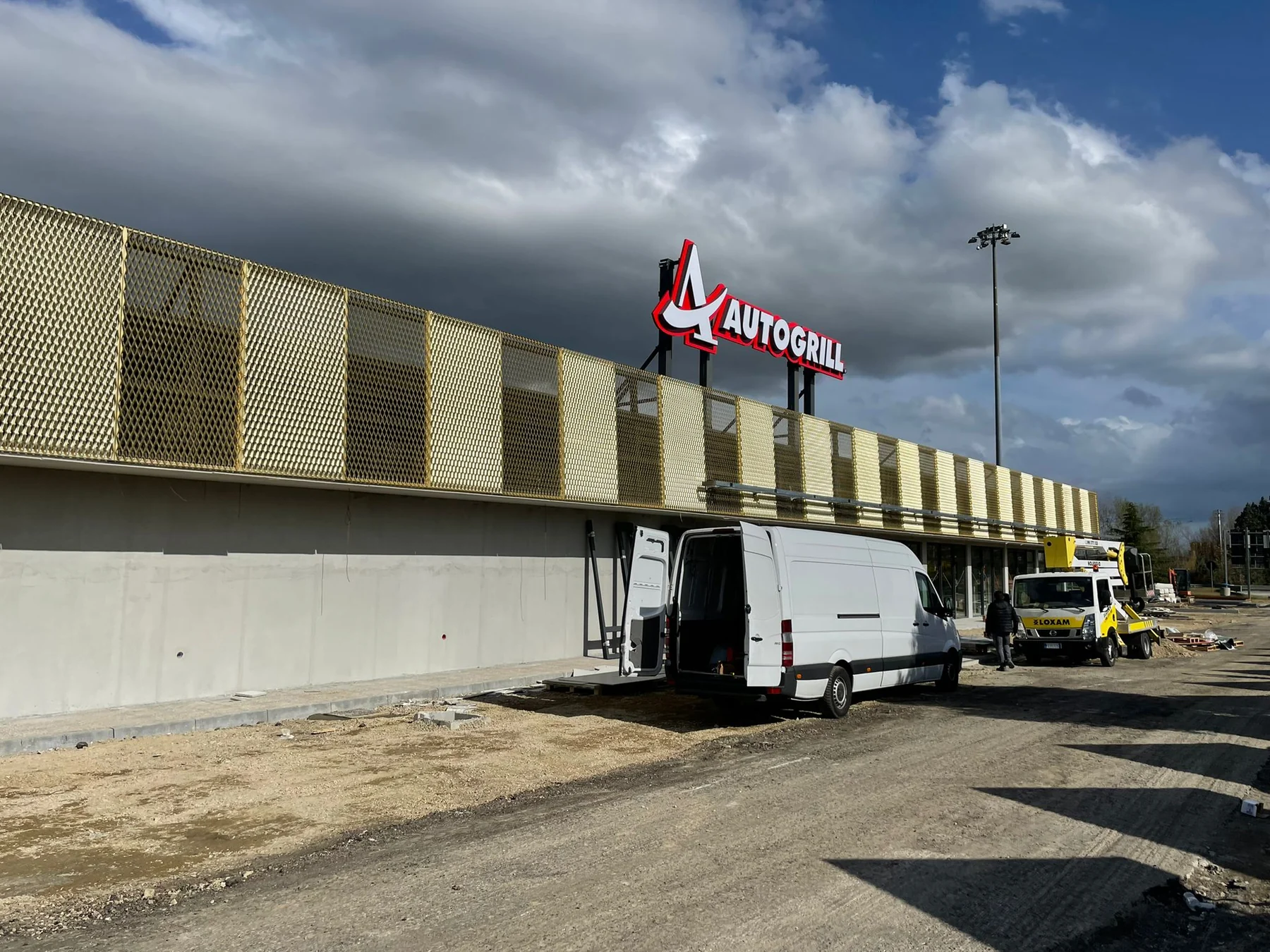 Vista ampia di Autogrill Montepulciano con insegna, mezzi operativi e materiali di cantiere.
