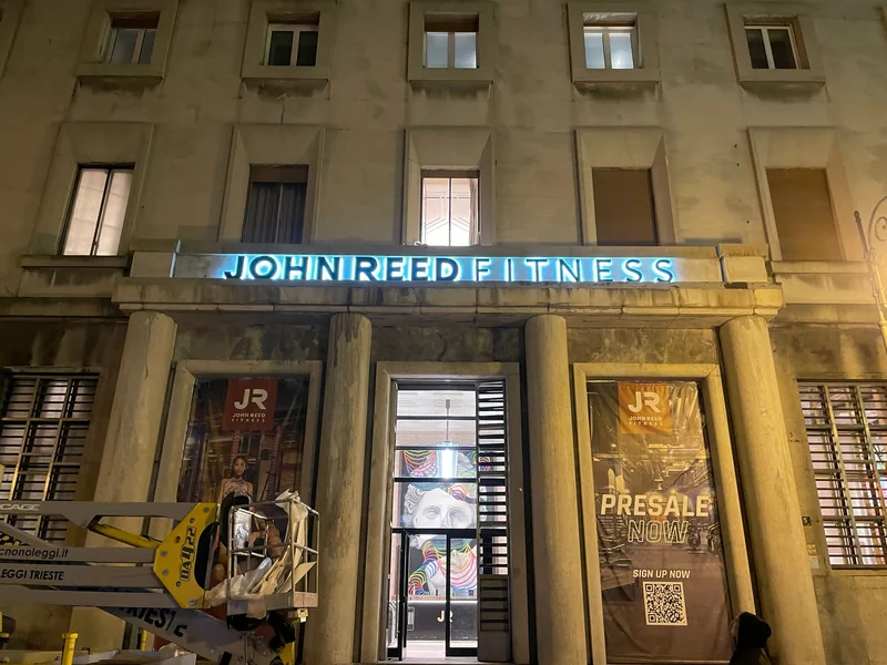 Vista frontale notturna dell’insegna azzurra John Reed Fitness a Trieste sopra l’ingresso, con una piattaforma di lavoro.
