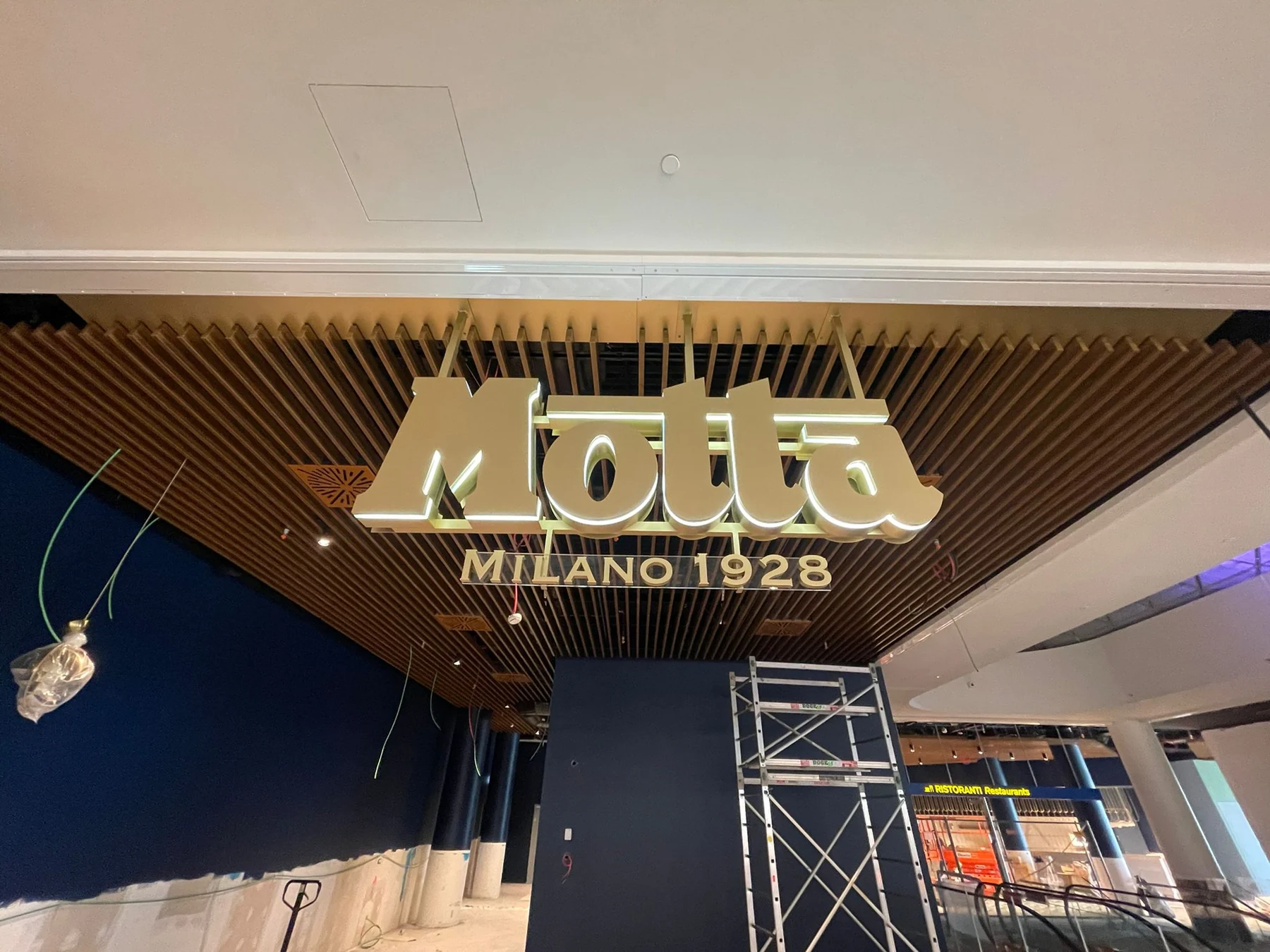 Vista frontale dell’insegna sospesa Motta Milano 1928, con attrezzature di cantiere ancora presenti.